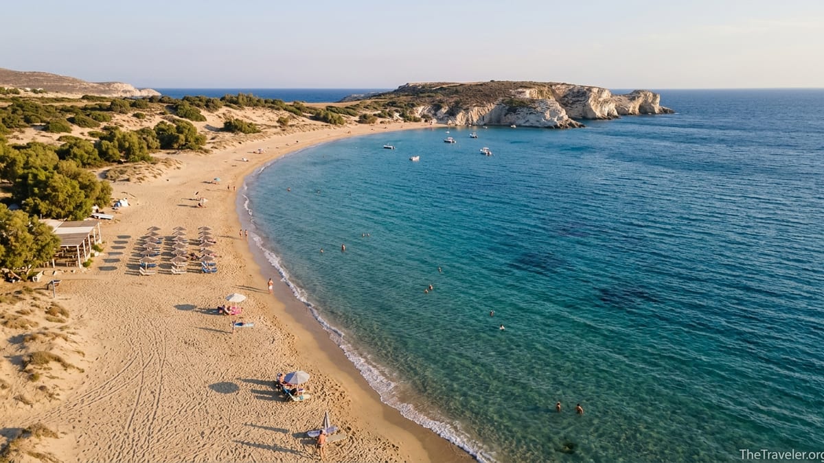 Aerial view of a Greek island beach with golden sand, turquoise water and cliffs at golden hour.