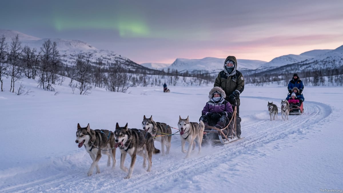 Couple and family on husky sleds crossing a snowy Arctic valley at twilight with mountains in the background.