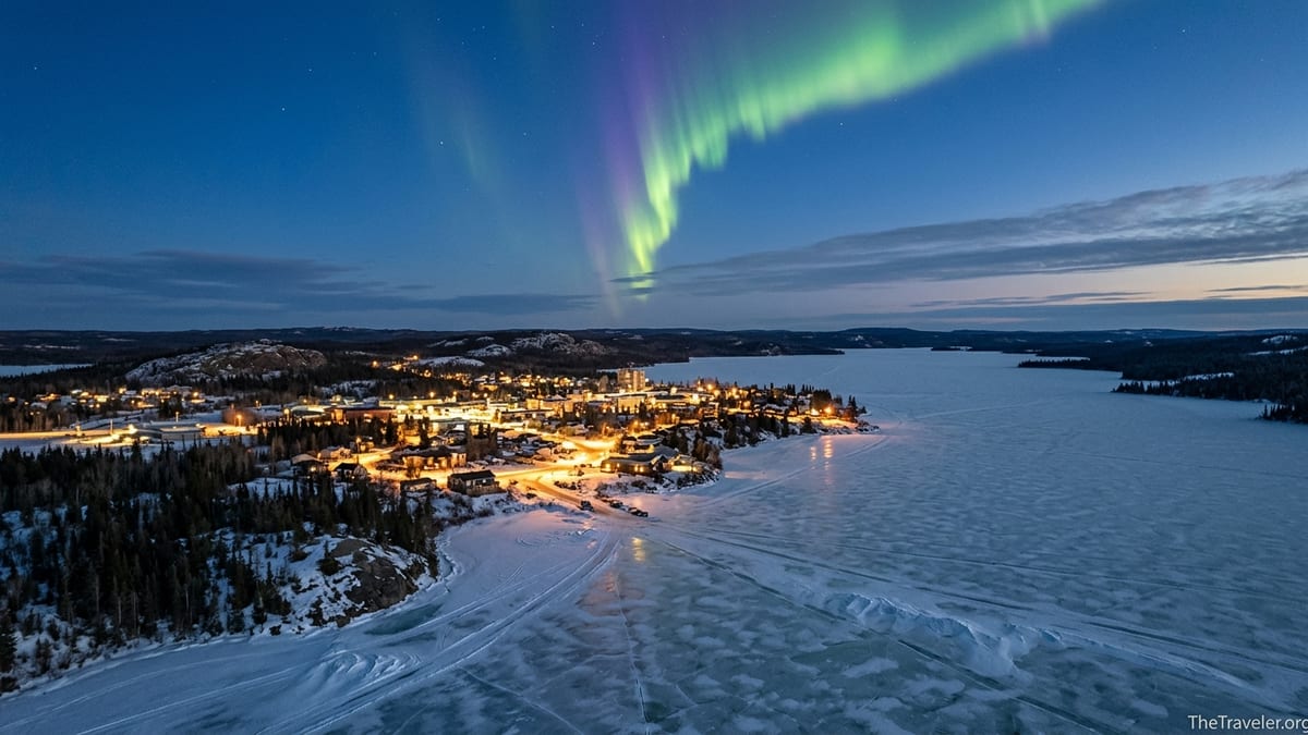 A northern Canadian town beside a frozen lake under bright aurora in a twilight sky.