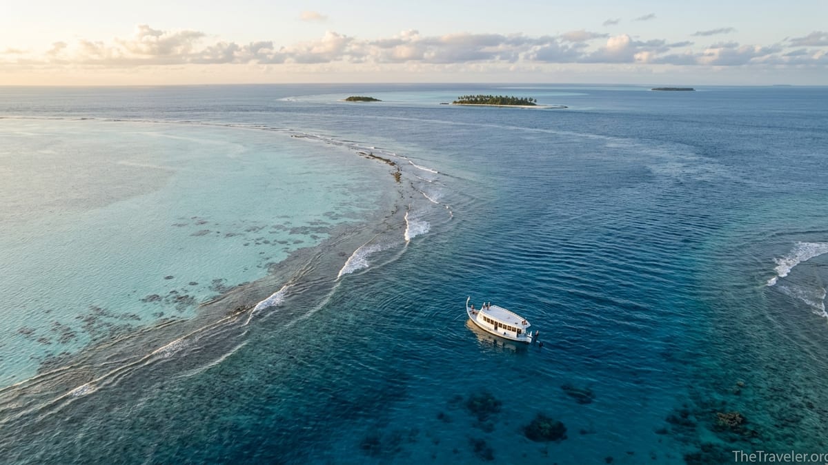 Aerial view of a Maldivian channel with a lone dive boat on clear turquoise water at sunrise.