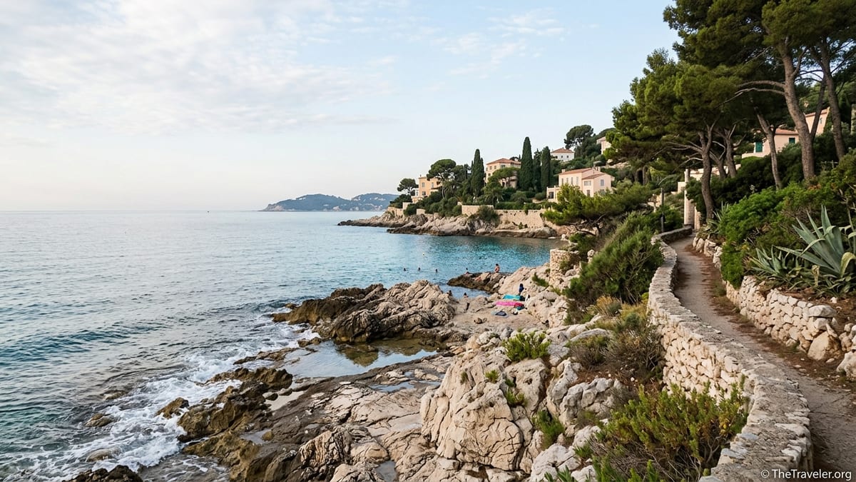 Rocky coastal path above a calm turquoise sea on Cap-Ferrat at sunrise, with pines and distant villas.