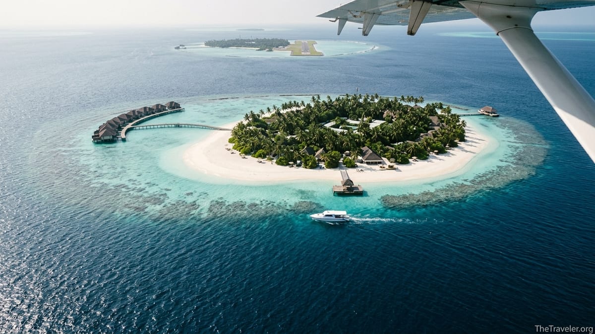 Aerial view of a Maldives resort island with overwater villas and a speedboat approaching through a turquoise lagoon.