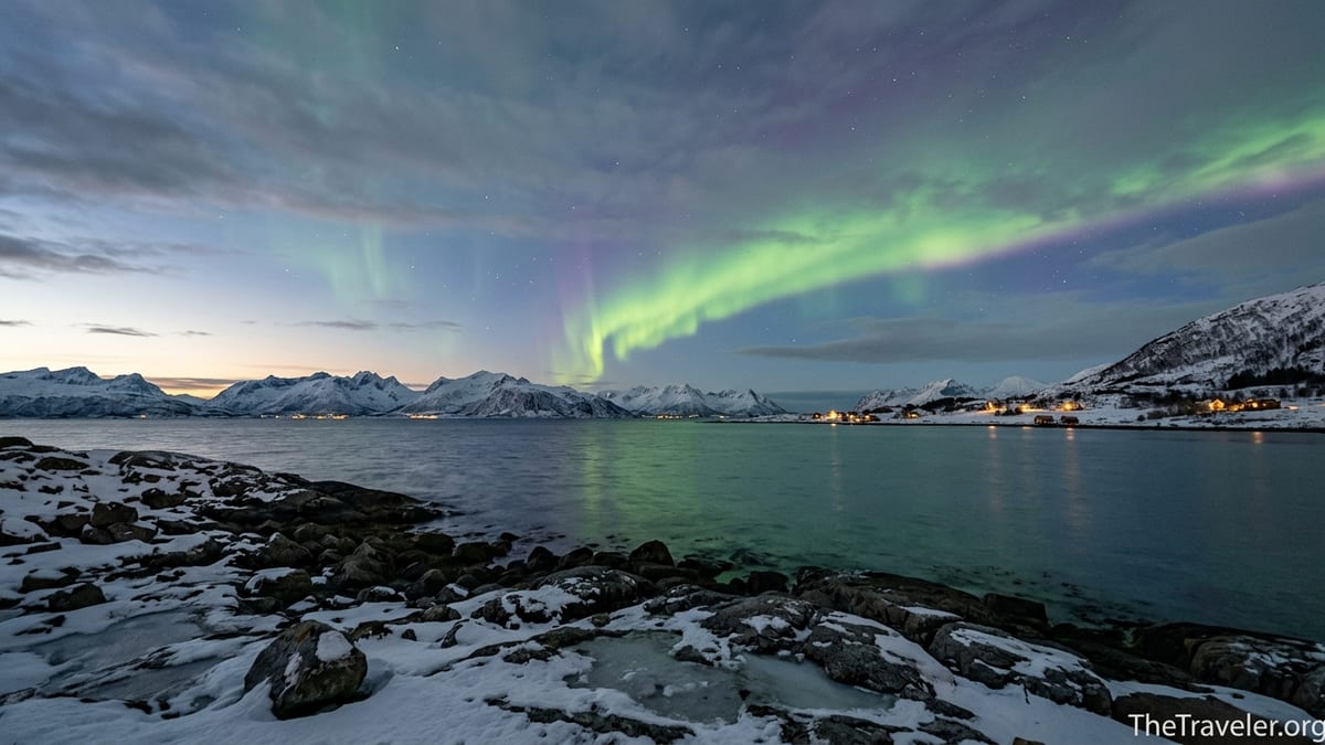 Green aurora stretching over snowy Norwegian fjord and mountains at dusk.