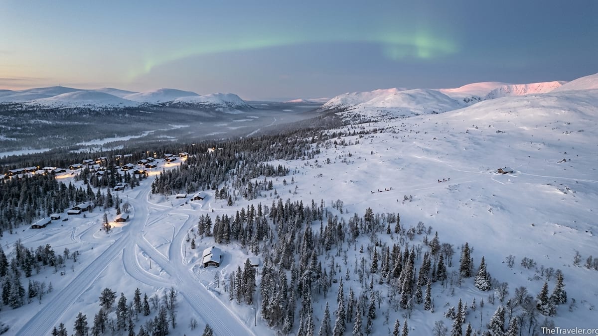 Aerial view of snowy Lapland fells showing ski tracks near cabins and vast untouched wilderness under northern lights.
