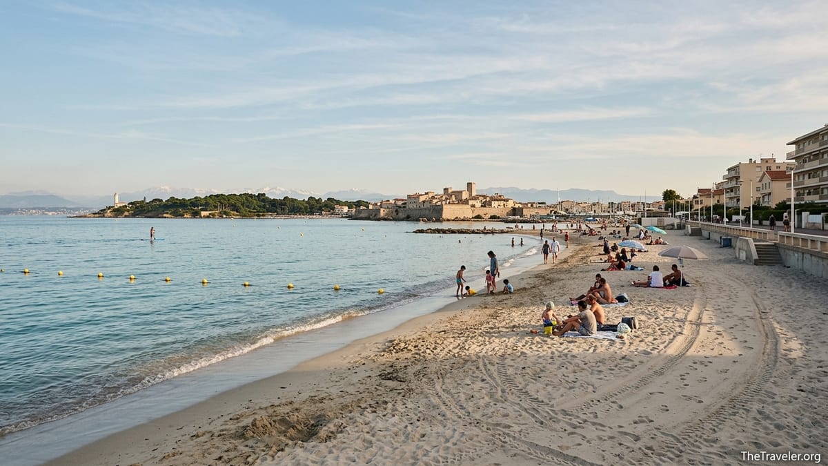 Late afternoon view along sandy Plage du Ponteil in Antibes with calm shallow sea and distant old town ramparts.