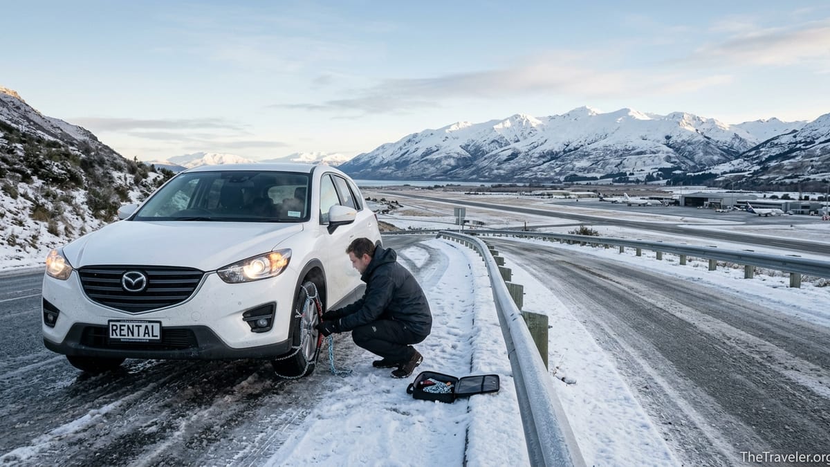 Driver fitting snow chains to a rental SUV near Queenstown Airport on an icy winter road.