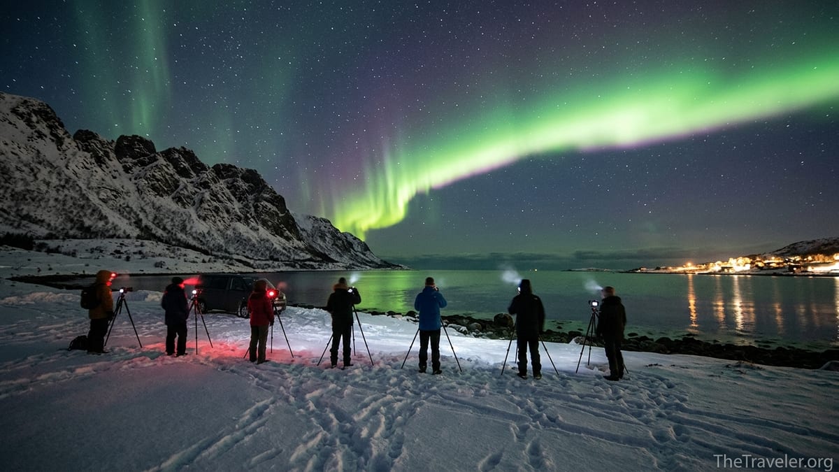 Small group of photographers under bright northern lights on a snowy Norwegian fjord.
