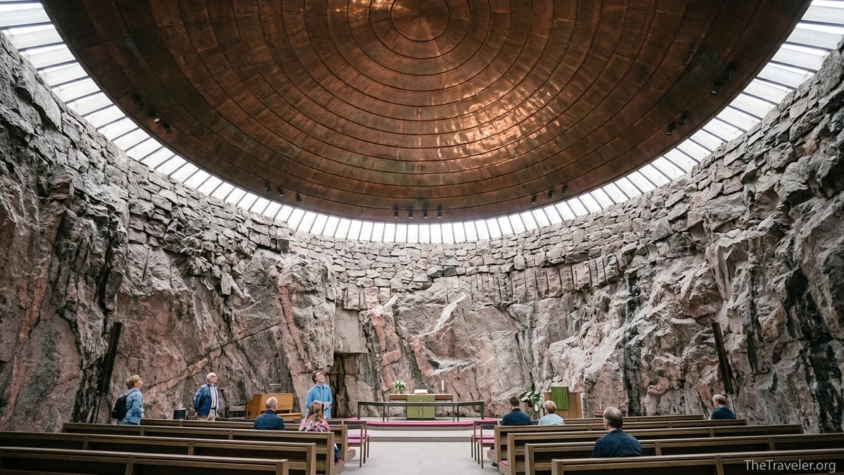 Interior of Temppeliaukio Church in Helsinki with rock walls, copper dome and visitors seated on wooden pews.