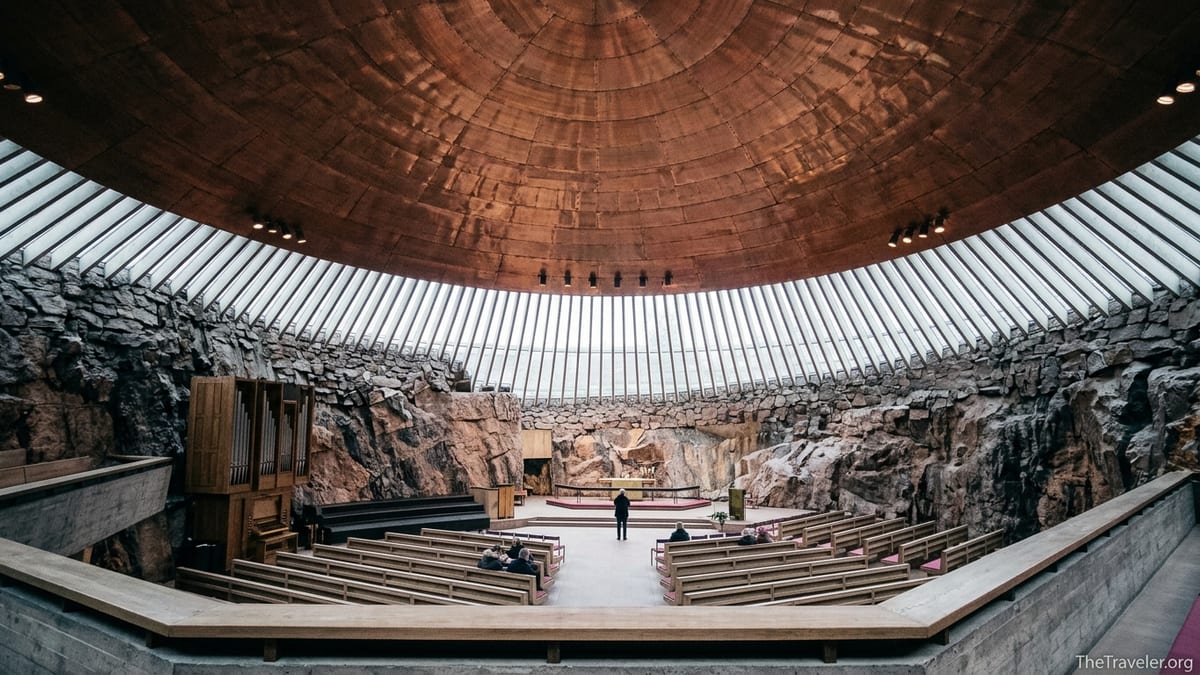 Interior of Temppeliaukio Church showing copper dome, rock walls and visitors sitting in wooden pews.