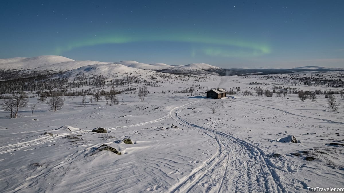 Snowy fells, ski trail and a small hut under northern lights in Urho Kekkonen National Park.