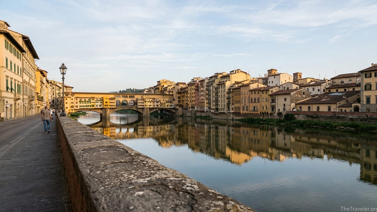 Sunrise view along the Arno River in Florence with Ponte Vecchio reflected in calm water.